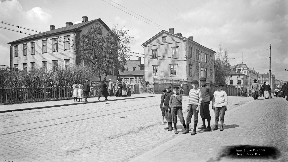 Näkymä Helsingin Siltasaarenkadulle. Etualalla kuuden nuoren pojan ryhmä katsoo kameraan. Kadun toisella puolella taustalla näkyy kaksi kerroksista puutaloa. Kadun yläpuolella risteilee ratikka ja puhelinkaapeleita. Kadun toisella puolella kulkee lisää ihmisiä.