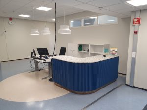 Etualalla näkyy puupaneloitu kaareva palvelutiski, jonka päällä on kivestä tehty pöytälevy. Tiskin vieressä on sähkökäyttöinen työpöytä, jolla on kaksi tietokonenäyttöä. Takaseinällä on avohylly.