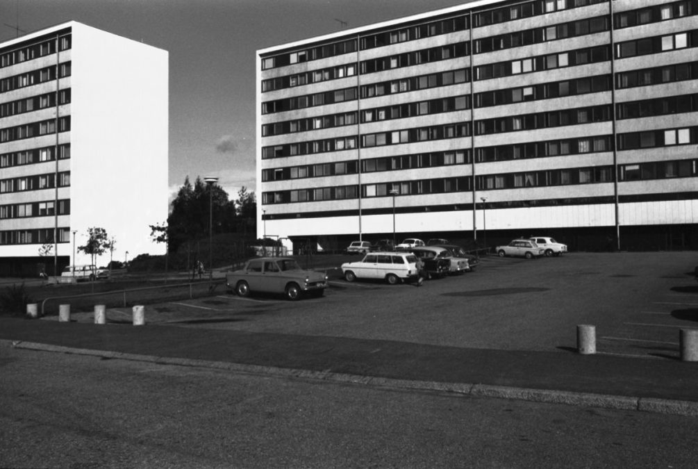 Taustalla neliskanttinen ja minimalistinen 1960-1970-lukujen taitteen kerrostalo. Etualalla parkkipaikka ja autoja. 