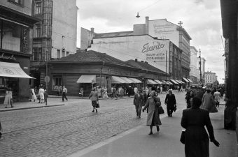 Kuvassa näkyy Aleksanterinkatu 9 ja Kluuvikatu 5 risteys. Kadulla kävelee paljon ihmisiä lämpimissä vaatteissa, mutta kadulla ei näy lunta. Kauempana kadulla näkyy yksi kuorma-auto