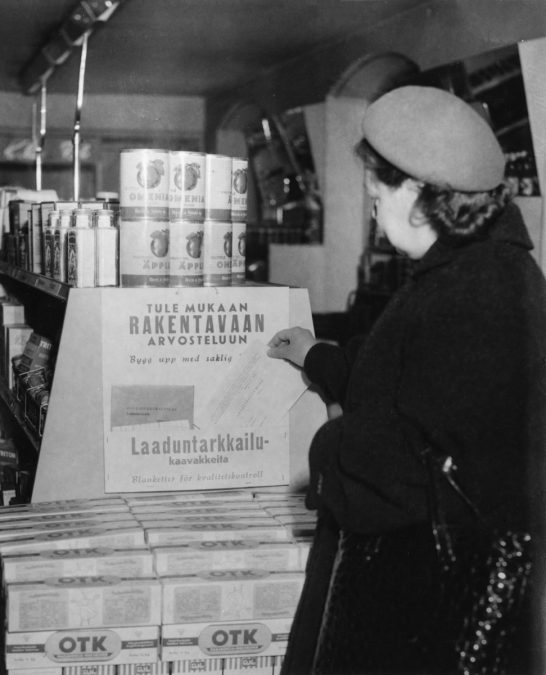 Turkiksiin ja baskeriin pukeutunut naishenkilö poimii OTK:n laaduntarkkailukaavakkeen. Kaavakkeen yläpuolella on teksti "Tule mukaan rakentavaan arvoteluun".