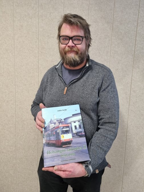 Keski-ikäinen parrakas ja silmälasipäinen mieshenkilö hymyilee kameralle kädessään kirja, jonka kannessa on vanha raitiotievaunu
